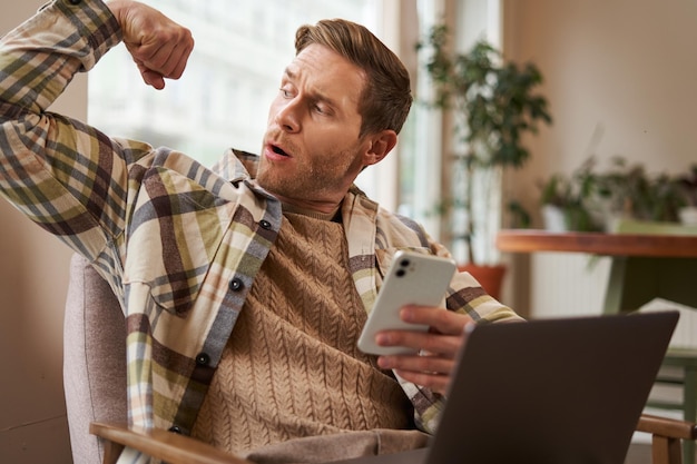 Foto gratuita retrato de un joven atlético sentado en un café con una computadora portátil y un teléfono inteligente que muestra sus bíceps flexionándose