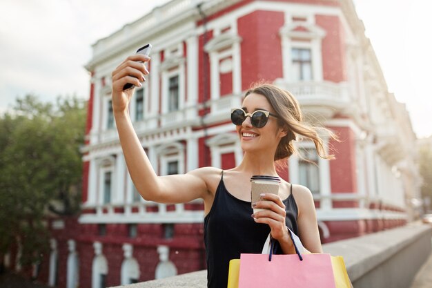 Retrato de joven atractiva chica caucásica femenina con cabello oscuro en gafas color canela y vestido negro sonriendo brillantemente tomando fotos frente a hermoso edificio rojo, bebiendo café, sosteniendo bolsas.