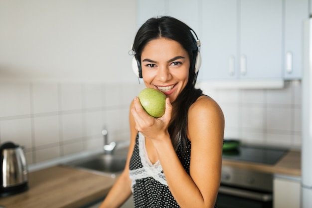 Retrato de joven atractiva cocinando en la cocina por la mañana, comiendo manzana, sonriendo, ama de casa feliz positiva, estilo de vida saludable, escuchando música en auriculares, riendo, dientes blancos de cerca