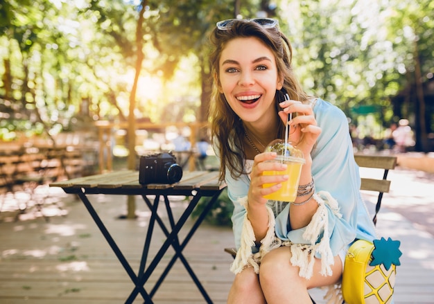 Retrato de joven atractiva y elegante mujer sentada en la cafetería, sonriendo sinceramente, bebiendo jugo sin problemas, estilo de vida saludable, estilo boho callejero, accesorios de moda, riendo, emoción feliz, soleado