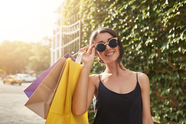 Retrato de joven y atractiva mujer europea de pelo castaño en gafas de sol y ropa negra sonriendo a la cámara, sosteniendo una gran cantidad de bolsas de la compra después de comprar regalos para amigos.