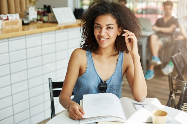 Retrato de joven atractiva mujer independiente de piel negra con cabello oscuro ondulado en camisa azul de moda cmiling brillantemente, mirando a la cámara con expresión relajada, sentado en el café sh