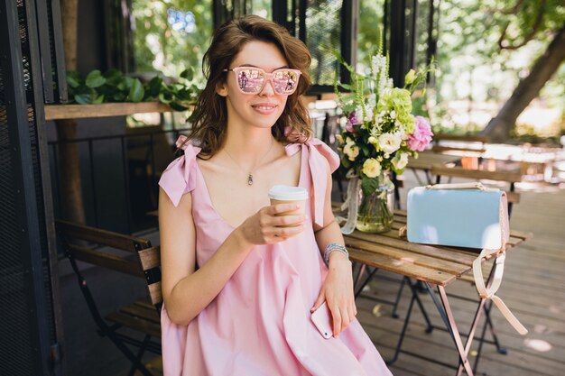 Retrato de joven atractiva mujer sentada en la cafetería en traje de moda de verano, vestido de algodón rosa, gafas de sol, sonriendo, tomando café, accesorios con estilo, ropa relajante y de moda