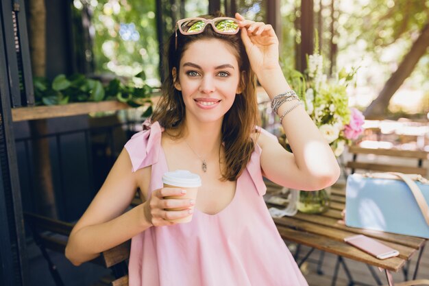 Retrato de joven atractiva mujer sentada en la cafetería en traje de moda de verano, vestido de algodón rosa, gafas de sol, sonriendo, tomando café, accesorios con estilo, ropa relajante y de moda