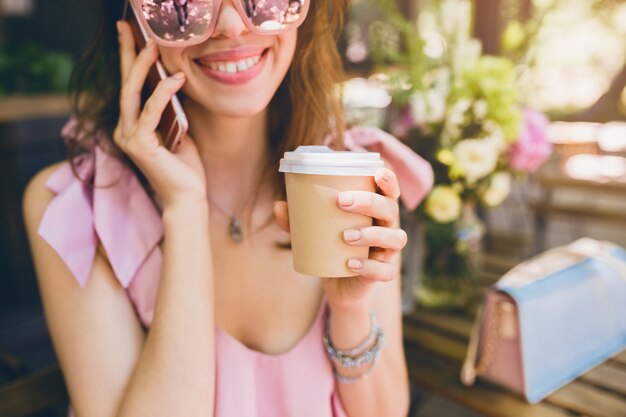 Retrato de joven atractiva mujer sentada en la cafetería, traje de moda de verano, vestido de algodón rosa, gafas de sol, sonriente, tomando café, accesorios elegantes, ropa de moda, hablando por teléfono