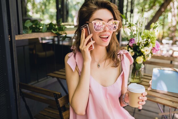 Retrato de joven atractiva mujer sentada en la cafetería, traje de moda de verano, vestido de algodón rosa, gafas de sol, sonriente, tomando café, accesorios elegantes, ropa de moda, hablando por teléfono