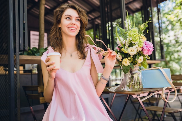 Retrato de joven atractiva mujer sentada en la cafetería, traje de moda de verano, vestido de algodón rosa, gafas de sol, sonriente, tomando café, accesorios elegantes, ropa de moda, humor feliz