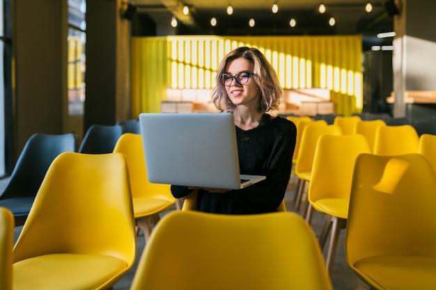 Retrato de joven atractiva mujer sentada en la sala de conferencias trabajando en la computadora portátil con gafas, estudiante aprendiendo en el aula con muchas sillas amarillas