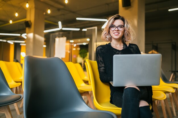 Retrato de joven atractiva mujer sentada en la sala de conferencias trabajando en la computadora portátil con gafas, estudiante aprendiendo en el aula con muchas sillas amarillas