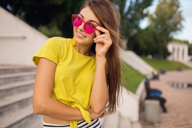 Retrato de joven atractiva mujer sonriente divirtiéndose en el parque de la ciudad, positivo, feliz, vistiendo top amarillo, aretes, gafas de sol rosadas, tendencia de moda de estilo veraniego, accesorios elegantes, colorido