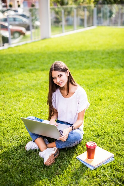 Retrato de joven atractiva, sentada sobre la hierba verde en el parque con las piernas cruzadas durante el día de verano mientras usa la computadora portátil