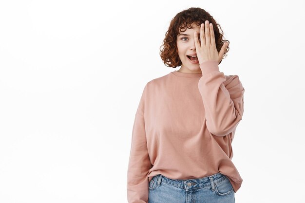 Retrato de una joven atractiva sonriendo, cubriendo la mitad de la cara y jadeando y luciendo emocionada, mirando algo asombroso, de pie con ropa informal sobre fondo blanco