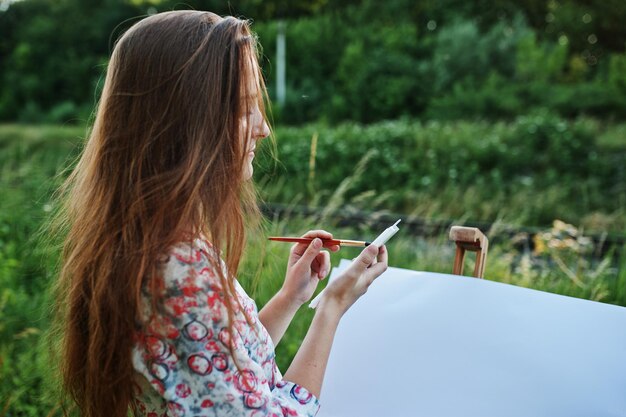 Retrato de una joven atractiva con un vestido largo pintando con acuarela en la naturaleza