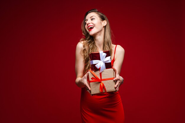 Retrato de joven atractiva en vestido de seda rojo sosteniendo pequeños regalos envueltos