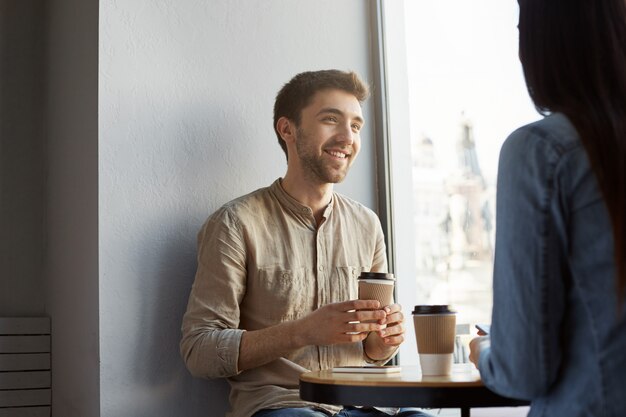 Retrato de joven atractivo sin afeitar con cabello oscuro, sonriendo, tomando café y escuchando historias de novia sobre el día duro en el trabajo. Estilo de vida, concepto de relación