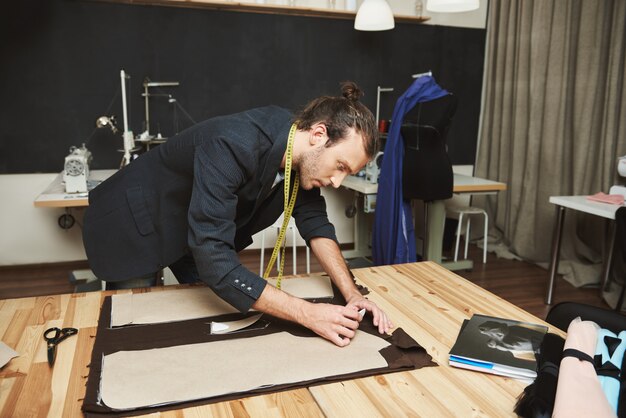 Retrato de un joven y atractivo diseñador de moda hispano en traje negro recortando partes de la chaqueta para la colección de invierno con expresión de la cara concentrada.