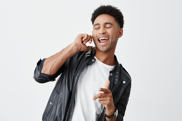 Retrato de joven atractivo estudiante americano de piel negra con cabello rizado en camiseta blanca y chaqueta de cuero cerrando los ojos, sosteniendo el dedo cerca de la oreja, cantando en voz alta en la fiesta.