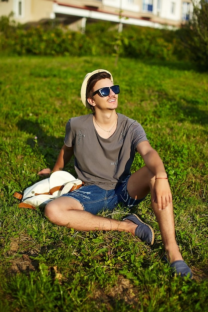 Retrato de joven atractivo hombre elegante moderno en ropa casual con sombrero en gafas sentado en el parque en la hierba verde