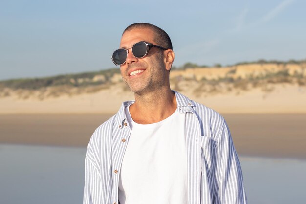 Retrato de joven atractivo en la playa. Modelo masculino con la cabeza rapada en gafas de sol sonriendo alegremente a la cámara. Retrato, vacaciones, concepto de belleza.