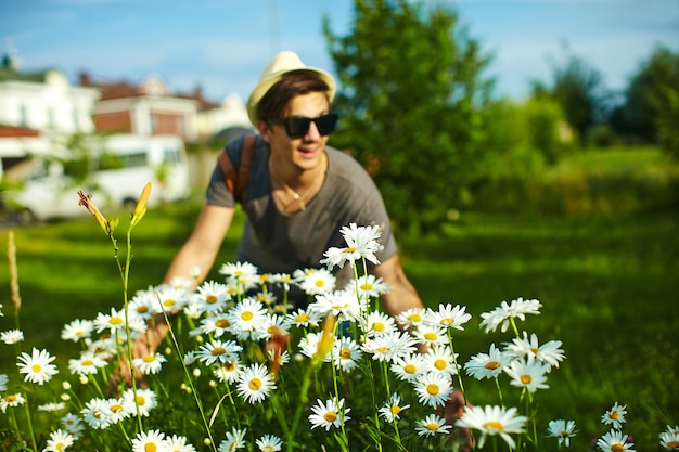 Retrato de joven atractivo sonriente hombre elegante moderno en ropa casual con sombrero en gafas en el parque con flores de colores brillantes en camomiles