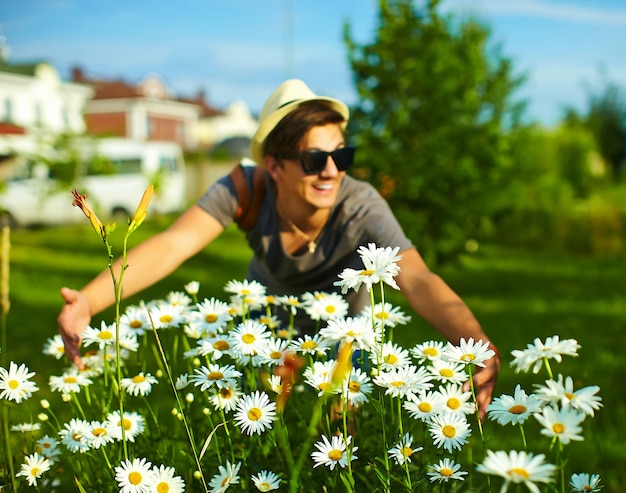 Retrato de joven atractivo sonriente hombre elegante moderno en ropa casual con sombrero en gafas en el parque con flores de colores brillantes en camomiles