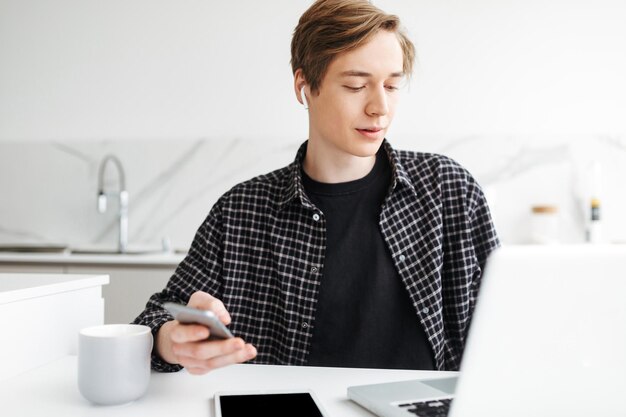 Retrato de un joven con auriculares sentado con un celular en la mano y trabajando en la cocina en casa aislado