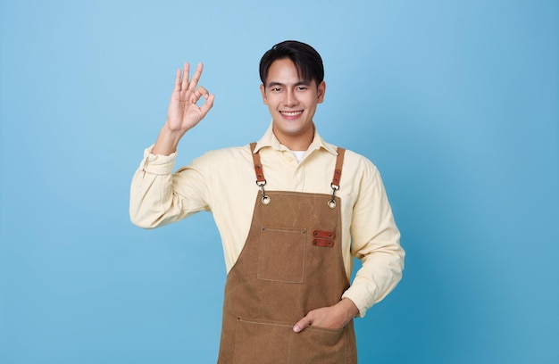 Retrato de un joven barista asiático con un delantal de pie gestando el signo OK