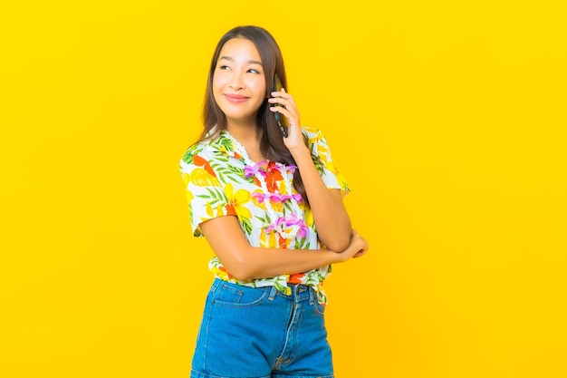 Retrato de joven y bella mujer asiática con camisa colorida con teléfono móvil inteligente en la pared amarilla
