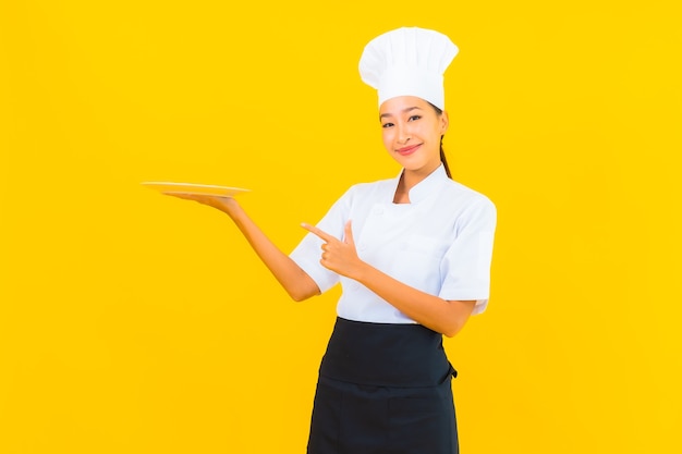 Retrato joven y bella mujer asiática chef con placa sobre fondo amarillo aislado