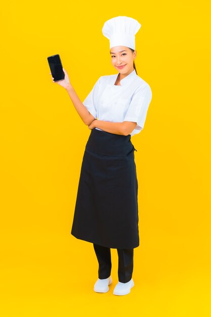 Retrato joven y bella mujer asiática chef con teléfono móvil inteligente sobre fondo amarillo aislado