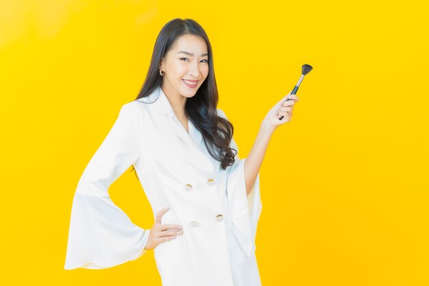 Retrato de joven y bella mujer asiática con maquillaje cosmético de pincel en pared amarilla
