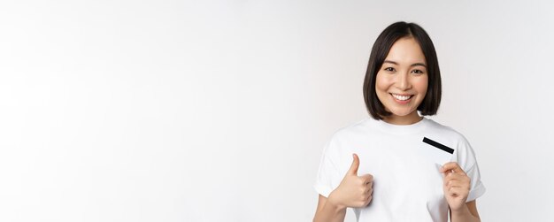 Retrato de una joven y bella mujer asiática moderna que muestra la tarjeta de crédito y los pulgares hacia arriba recomendando contacto