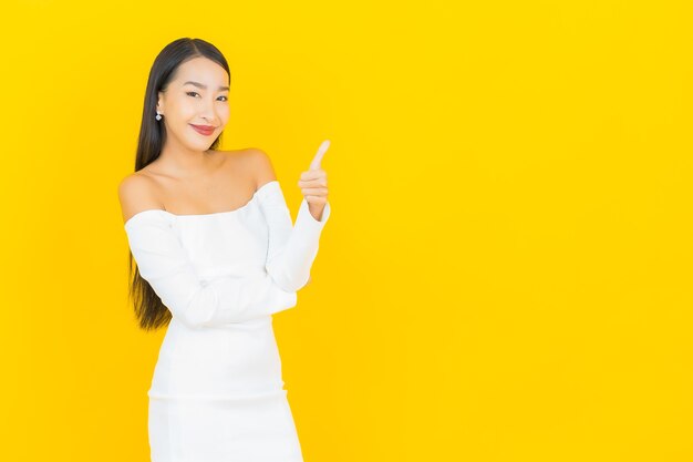 Retrato de joven y bella mujer asiática de negocios sonriendo y dando pulgar hacia arriba con vestido blanco en la pared amarilla