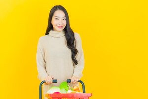 Foto gratuita retrato joven y bella mujer asiática sonríe con la cesta de la compra del supermercado en la pared amarilla