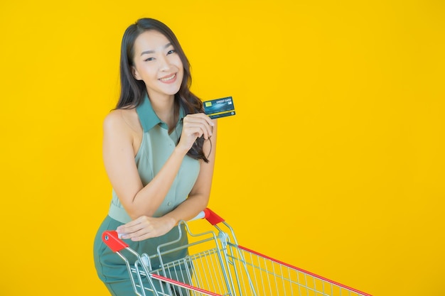 Retrato de joven y bella mujer asiática sonríe con la cesta de la compra del supermercado en la pared amarilla