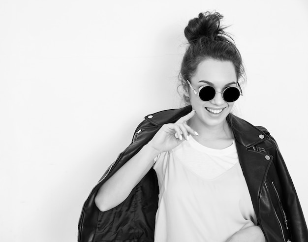 Retrato de joven y bella mujer morena modelo de niña con maquillaje desnudo vistiendo ropa de verano hipster motorista chaqueta de cuero en gafas de sol posando junto a la pared.