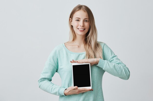 Retrato de joven bella mujer rubia sonriente sosteniendo y mostrando tableta digital en blanco