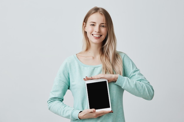 Retrato de joven bella mujer rubia sonriente sosteniendo y mostrando tableta digital en blanco