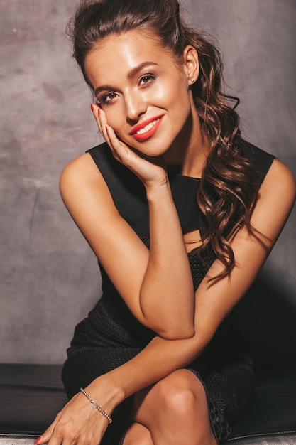 Retrato de joven bella mujer sonriente hipster en vestido de moda verano negro. Mujer despreocupada sexy posando junto a la pared. Modelo morena con maquillaje y peinado. Sentado en la silla