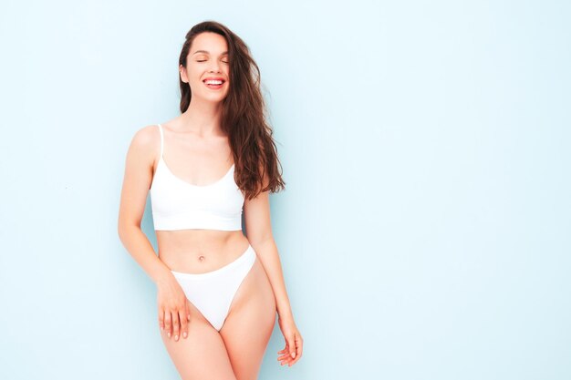 Retrato de joven bella mujer sonriente en lencería blanca. Sexy modelo alegre despreocupada en ropa interior posando junto a la pared azul claro en el estudio. Mujer positiva y feliz disfrutando de la mañana