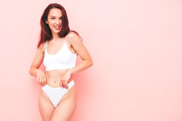 Retrato de joven bella mujer sonriente en lencería blanca. Sexy modelo alegre despreocupada en ropa interior posando junto a la pared rosa en el estudio. Mujer positiva y feliz disfrutando de la mañana