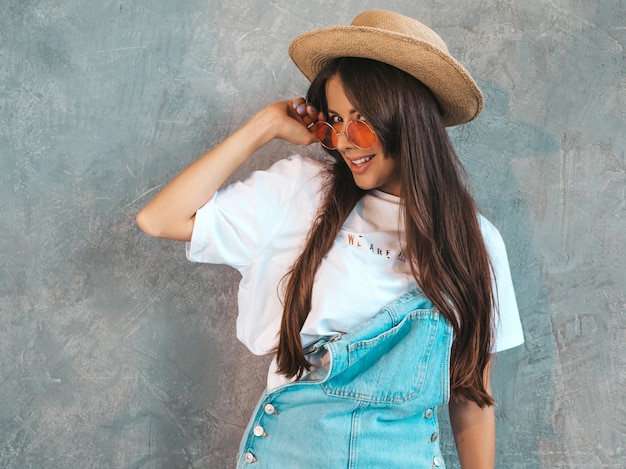 Retrato de joven bella mujer sonriente mirando. Chica de moda en ropa de verano casual monos y sombrero.