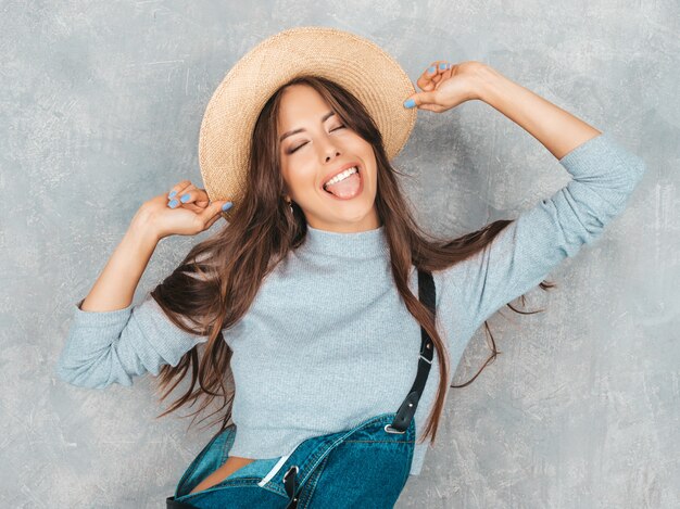 Retrato de joven bella mujer sonriente con los ojos cerrados. Chica de moda en ropa de verano casual monos y sombrero. y muestra lengua