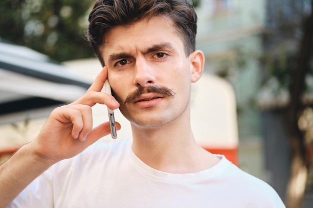 Retrato de un joven bigote disgustado con una camiseta blanca hablando por teléfono celular mientras mira cuidadosamente a la cámara en la calle de la ciudad