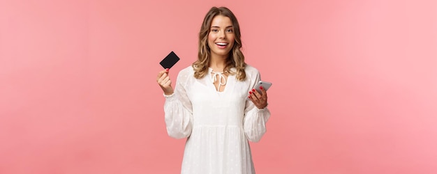 Retrato de una joven y bonita mujer rubia despreocupada que usa un teléfono móvil para pagar una compra en línea con una tarjeta de crédito y una cámara feliz sonriente que compra una aplicación de teléfono inteligente para descargar en Internet