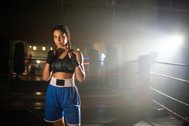 Retrato de una joven boxeadora posando para la cámara en el gimnasio. Hermosa chica en ropa deportiva de pie dentro del ring de boxeo mostrando sus puños en vendas y mirando a la cámara. Concepto de boxeo deportivo y femenino.