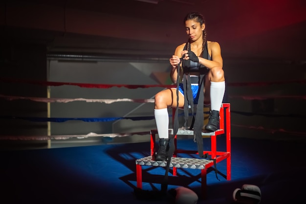 Retrato de una joven boxeadora preparando sus manos para la pelea. Hermosa chica sentada en las escaleras para el anillo, envolviendo cuidadosamente puños fuertes con vendas para el boxeo activo. Concepto de boxeo deportivo y femenino.