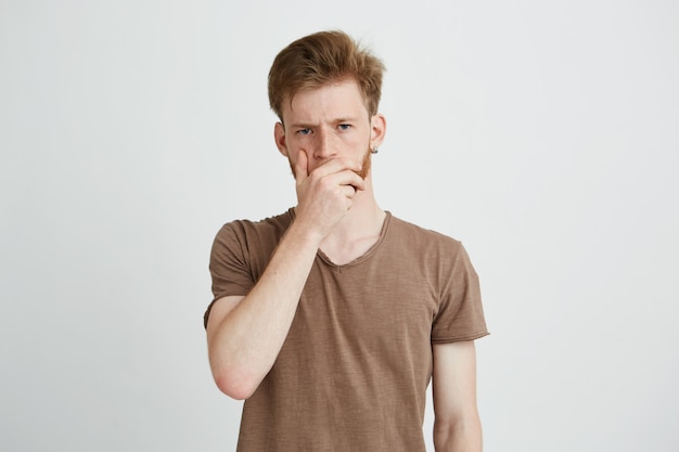 Retrato de joven brutal guapo con barba frunciendo el ceño la mano en la boca.