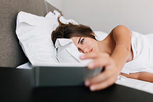 Retrato joven en cama en apartamento moderno por la mañana. Ella sostiene el teléfono y se ve cansada.