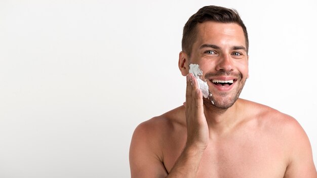 Retrato de joven sin camisa aplicando espuma de afeitar de pie contra la pared blanca